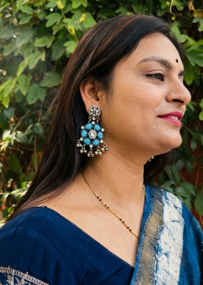 Woman wearing a blue saree with intricate patterns, standing against a green leafy background.