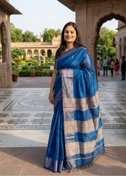 Woman in a blue saree standing in an outdoor setting with architectural elements and people in the background.