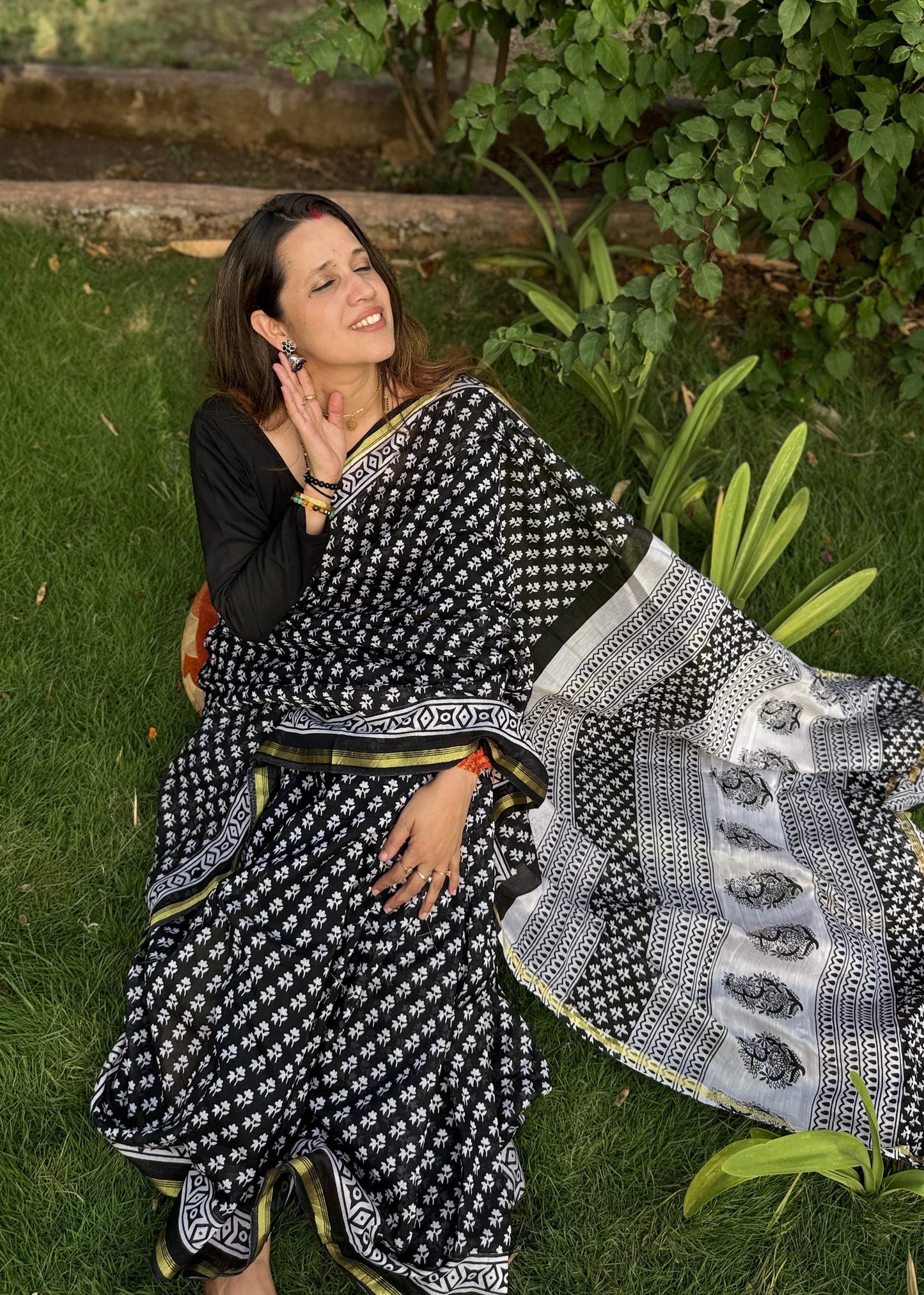 Woman in a black and white saree sitting on grass with greenery around