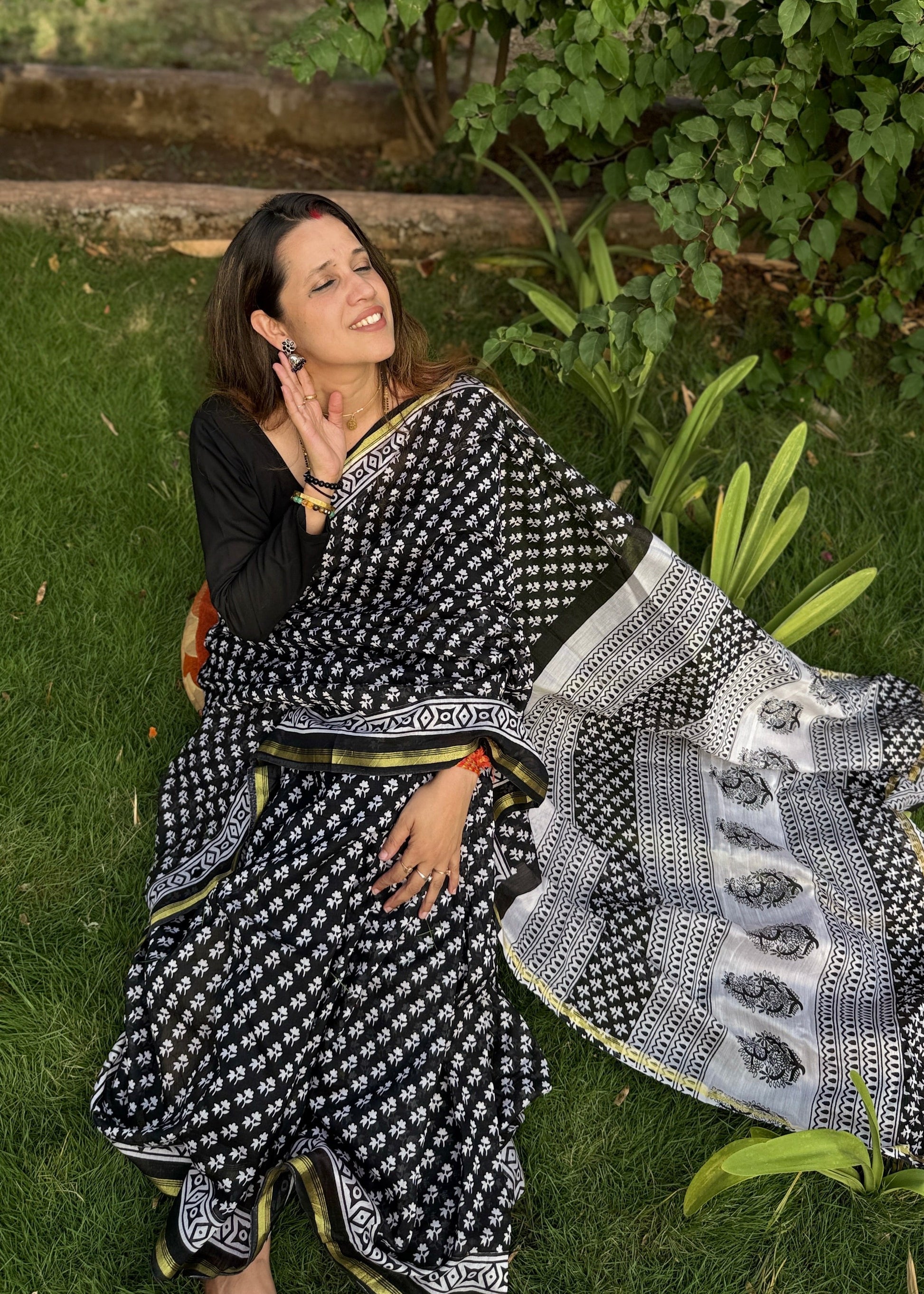 Woman in a black and white saree sitting on grass with greenery around