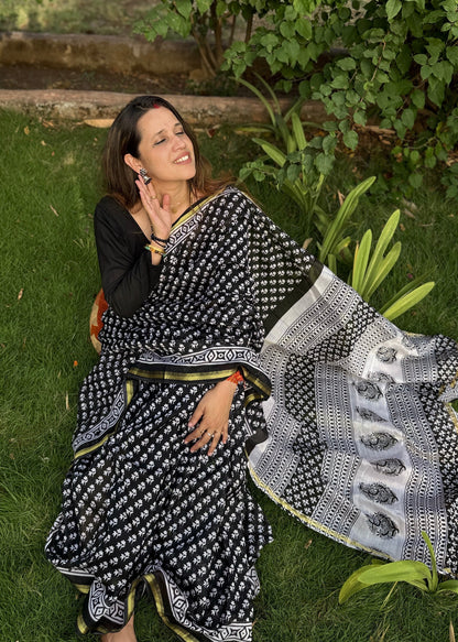 Woman in a black and white saree sitting on grass with greenery around