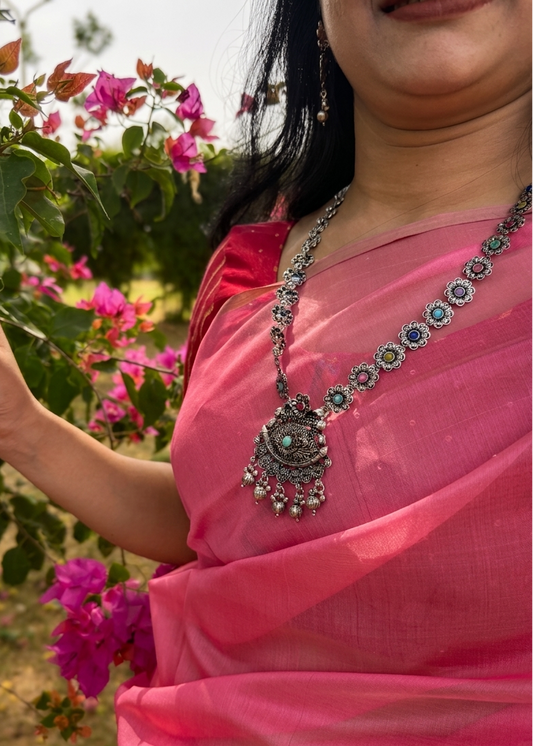 Woman wearing a pink saree with a decorative necklace, standing in front of pink flowers.