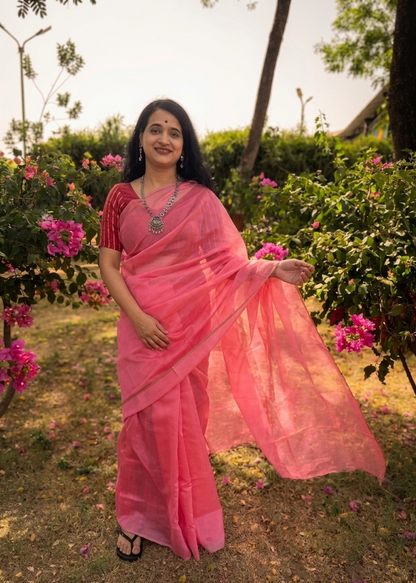Woman in a pink saree standing in a garden with flowers and greenery.