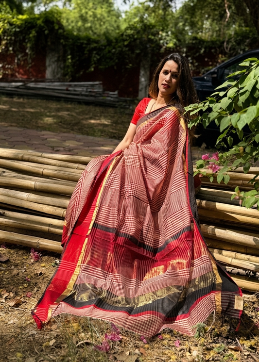 Woman in a red and black saree standing outdoors with greenery in the background