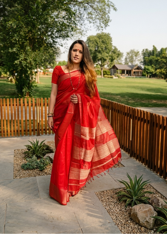 Woman in a red saree standing outdoors with greenery and a wooden fence in the background