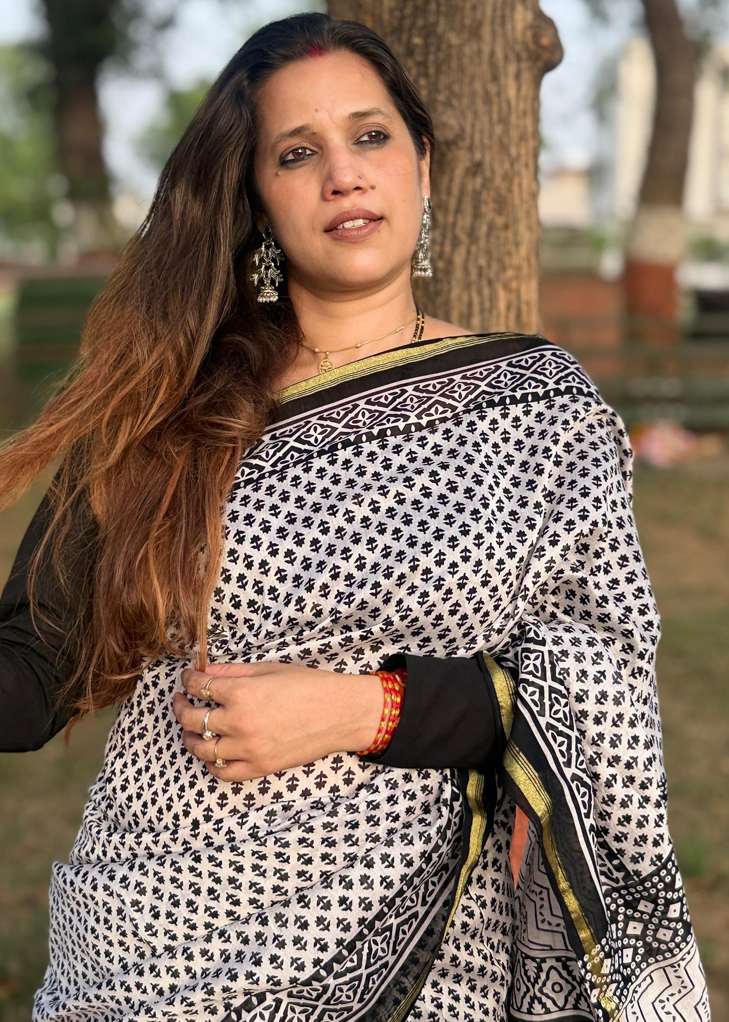 Woman wearing a black and white patterned saree standing outdoors with trees in the background