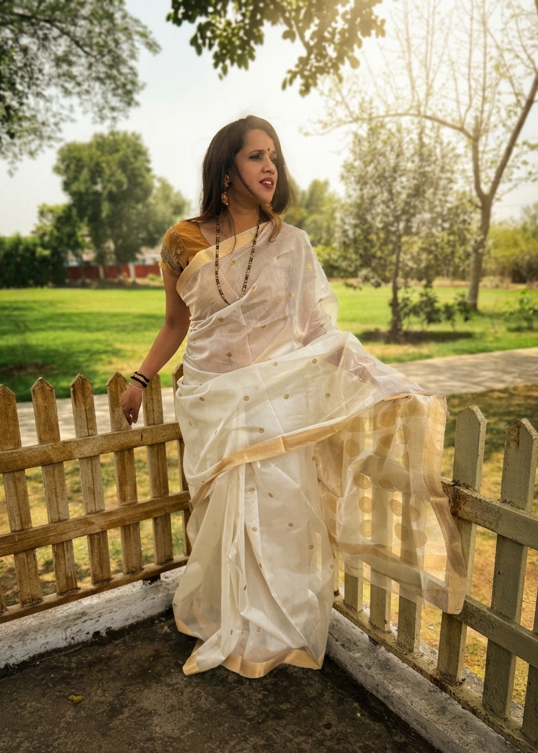 Woman in a white saree standing by a wooden fence with greenery in the background